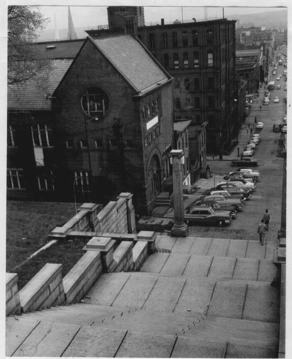 Photograph showing several students walking down the bottom of The Approach steps, with the old gymnasium building, located at the intersection of Broadway and Sixth Street, visible to the left (date unknown, possibly circa 1950s)