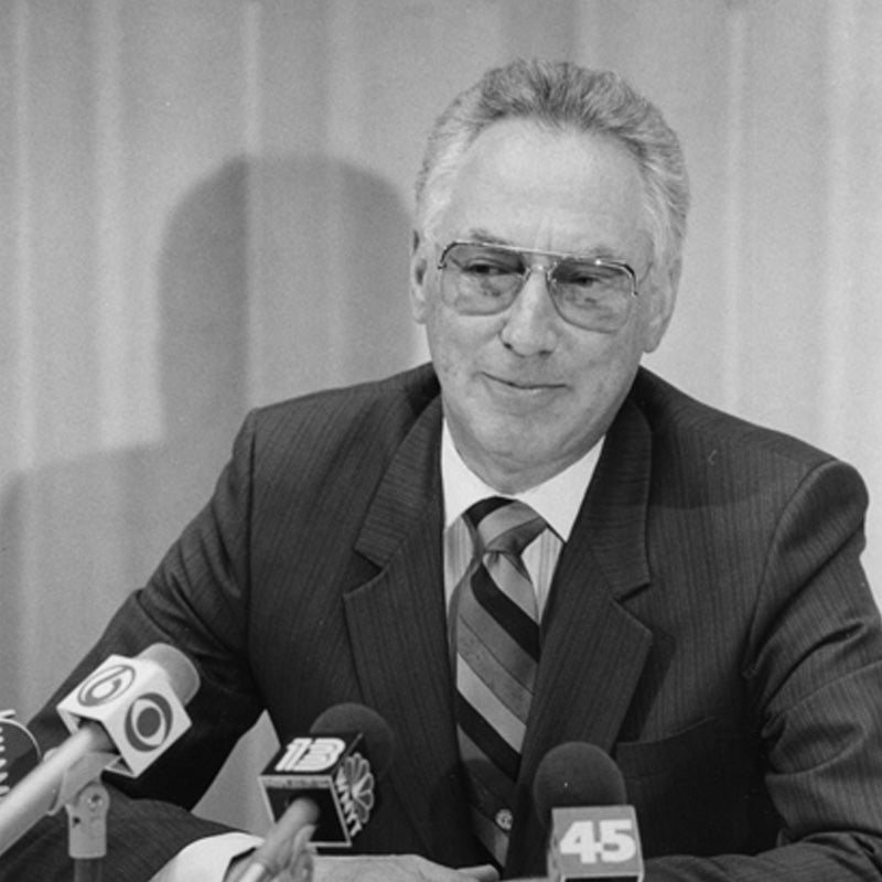 Black-and-white photograph of Stanley Landgraf, seated before several news channel microphones